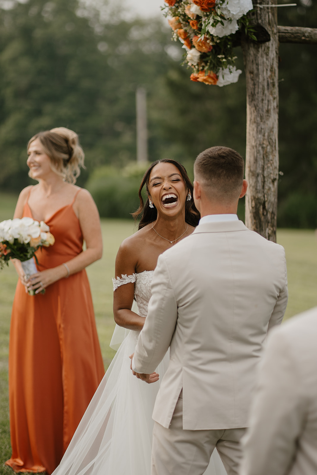 Bride laughs with the groom and a bridesmaid in orange beneath the floral ceremony arch.