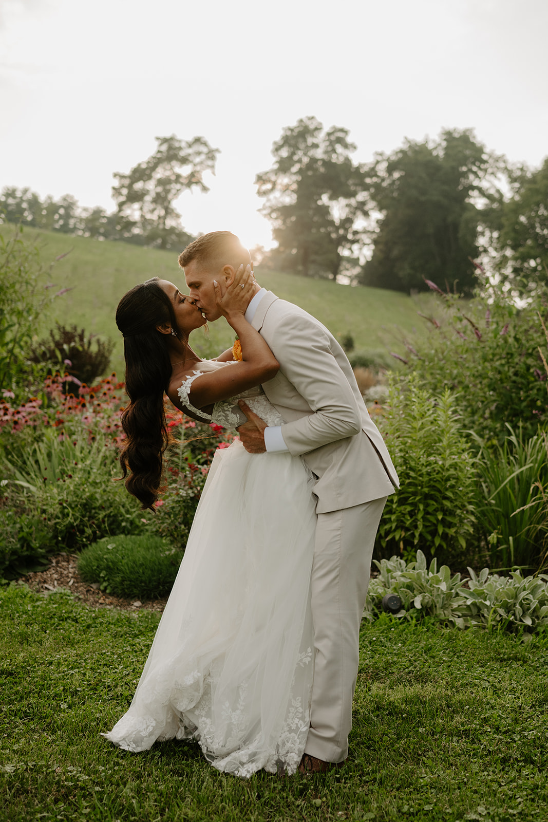 Groom dips the bride for a kiss beside the garden at golden hour.