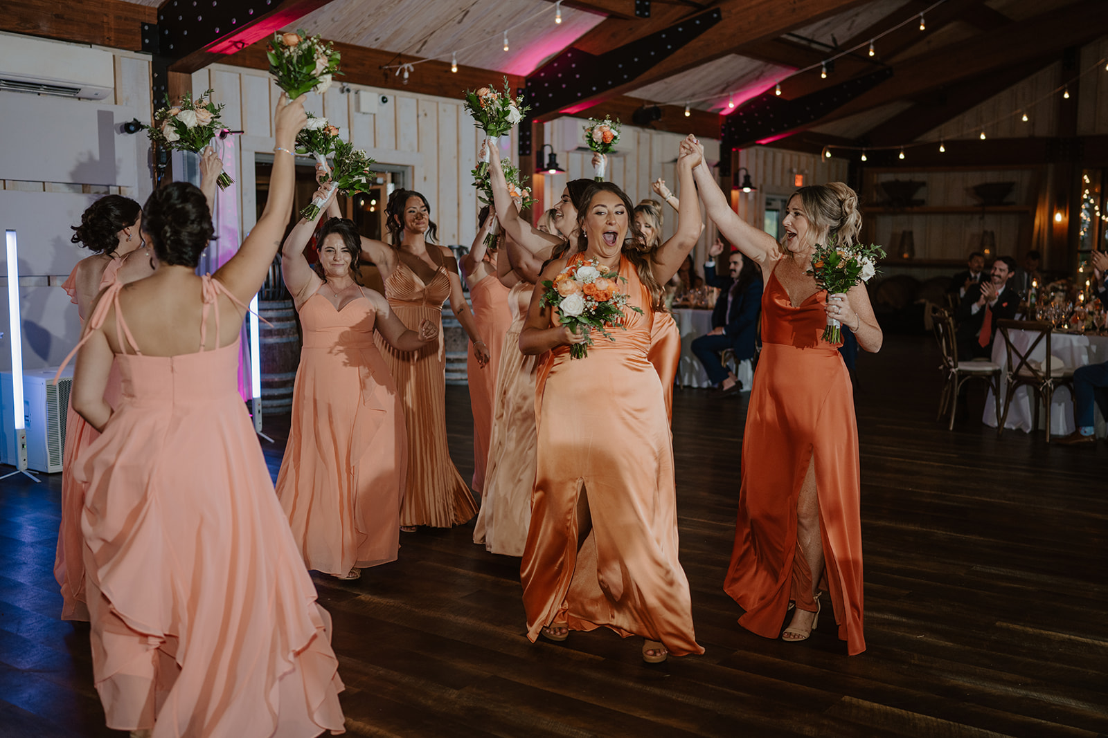 Bridesmaids dance into the reception with bouquets held high under string lights.