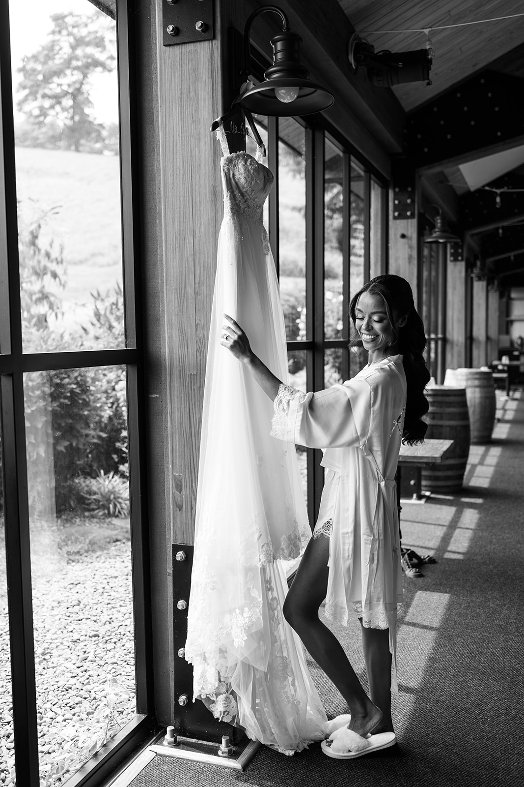 Black and white photo of the bride reaching for her gown hanging in a sunlit doorway.