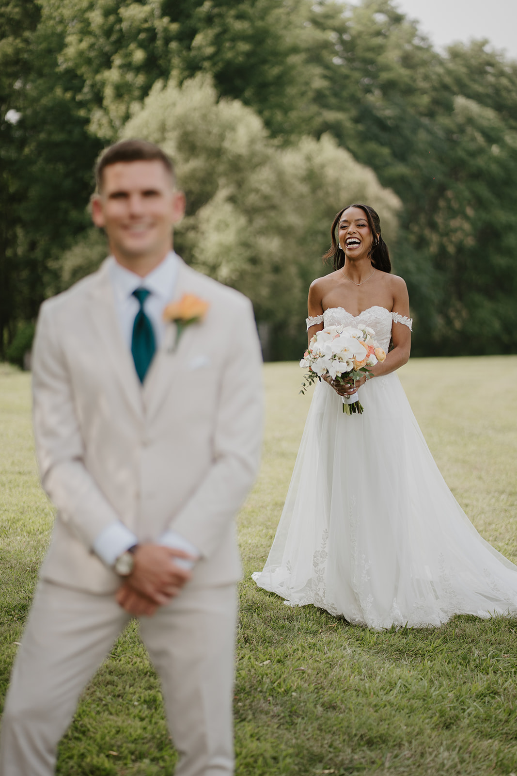 Groom stands in the foreground as the bride laughs behind him on the lawn.