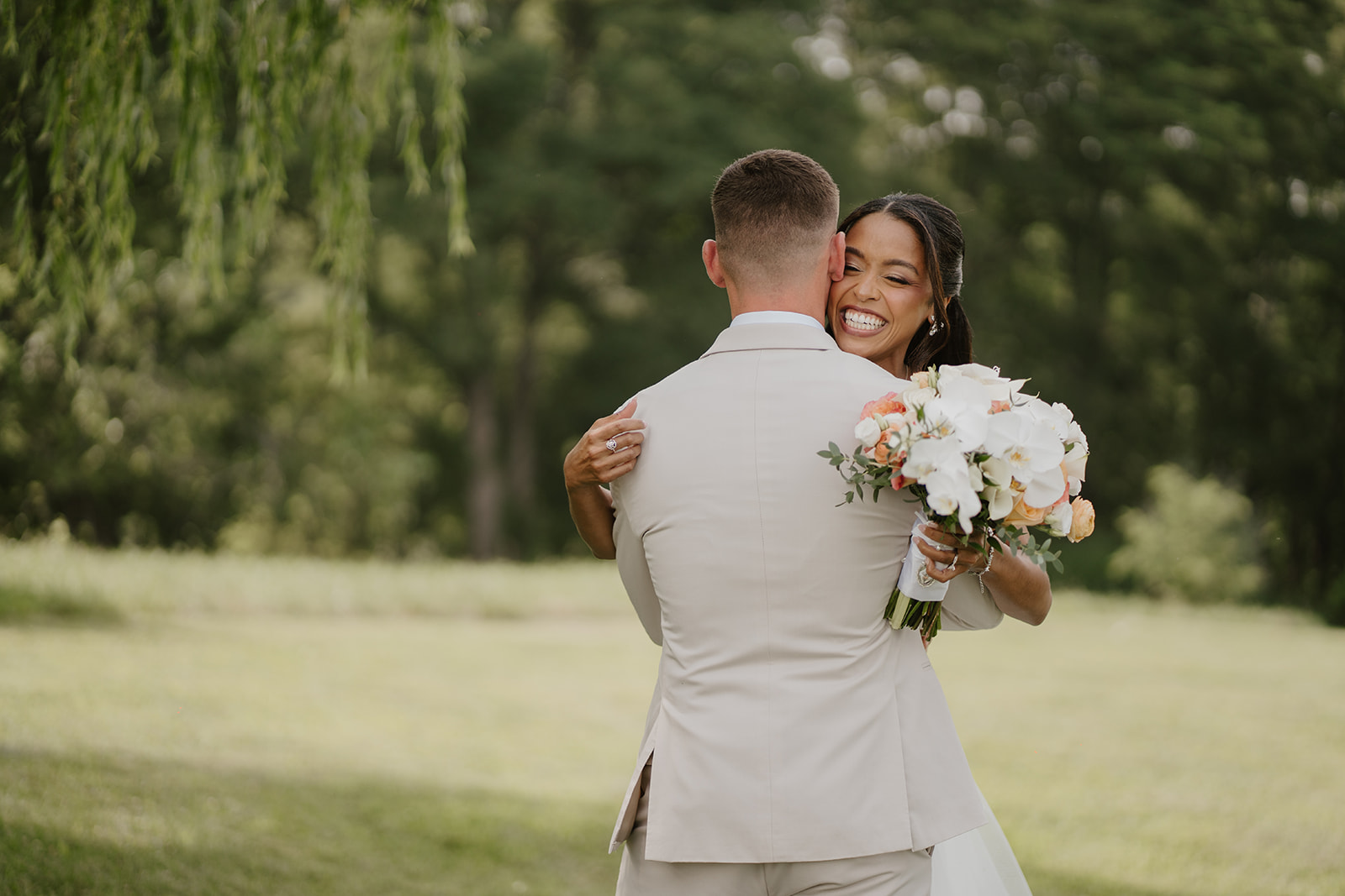 Bride hugs the groom from behind, her bouquet resting on his shoulder.