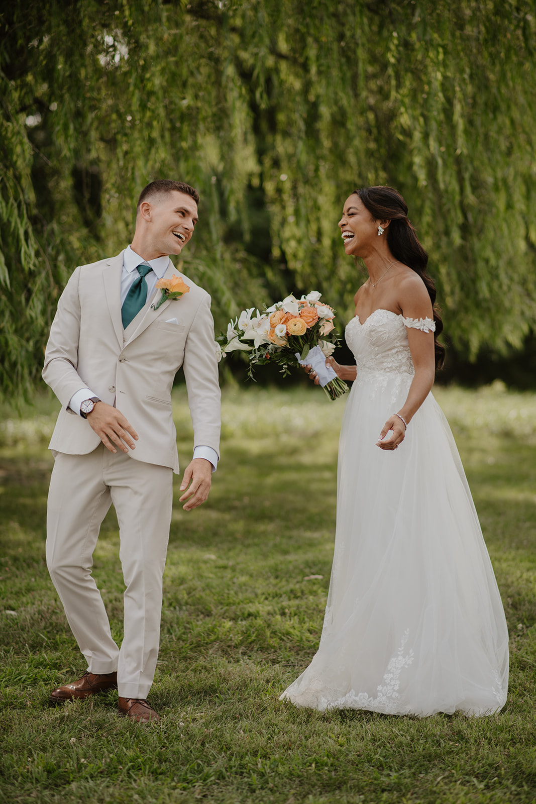 Bride and groom walk the lawn together, laughing beneath the willow branches.