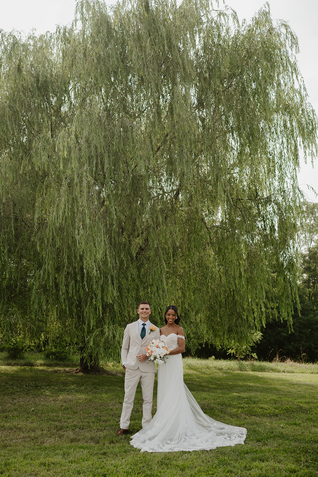 Bride and groom stand hand in hand on the lawn framed by the willow.