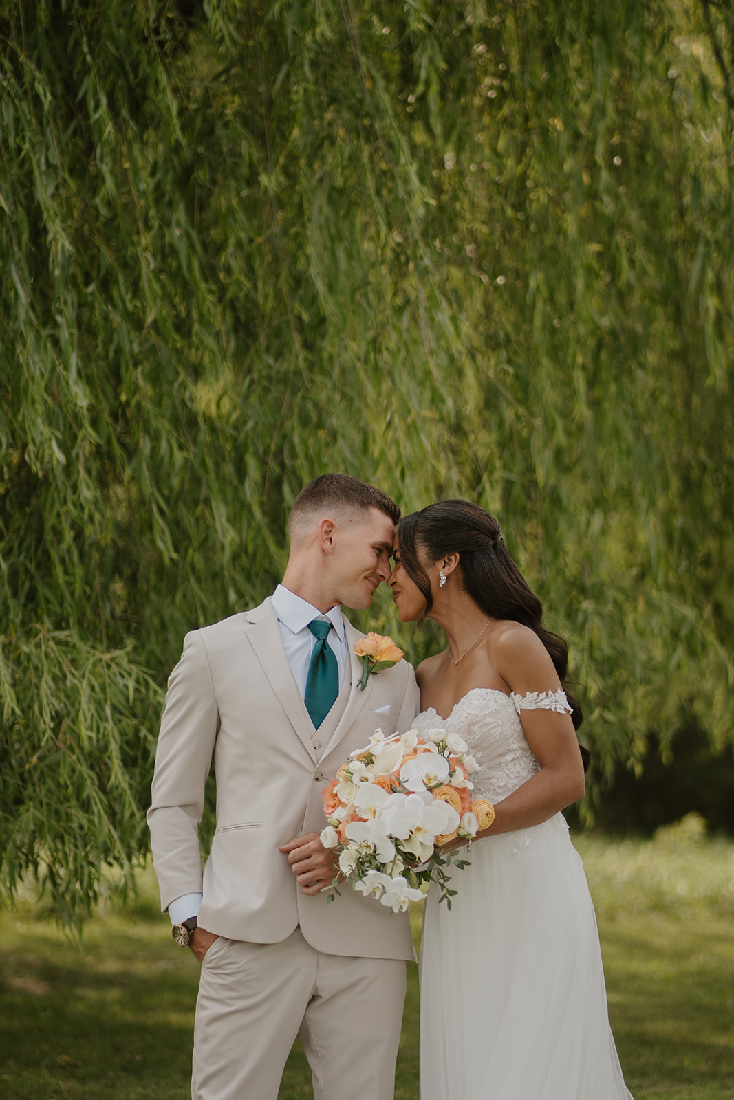 Bride and groom lean in close beneath the willow, her bouquet between them.