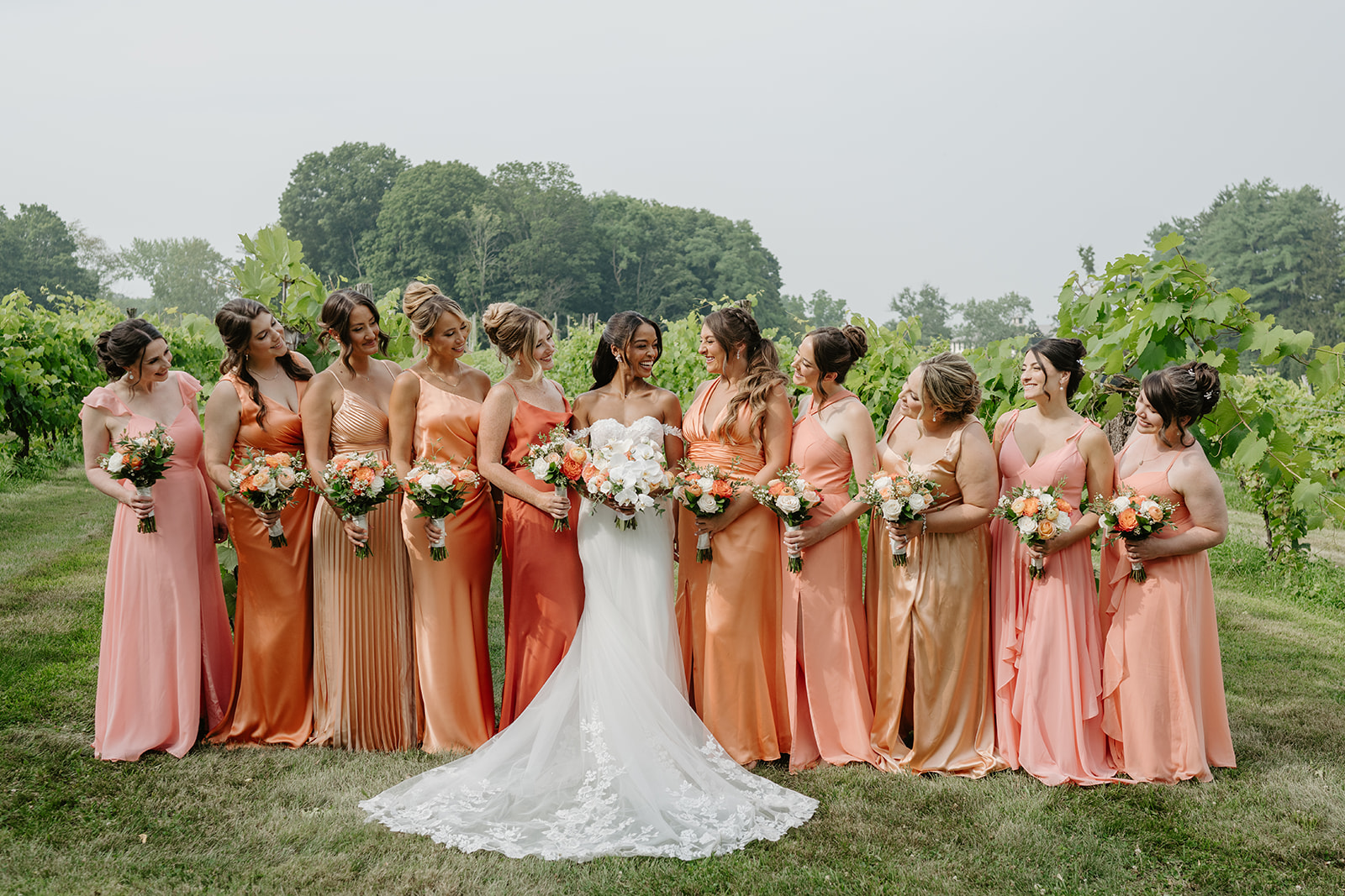 Bride stands surrounded by her bridesmaids in coral gowns beside the vineyard.