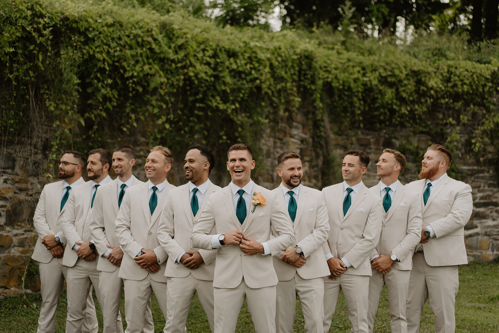 Groomsmen laugh together in a row before the ivy-covered wall.