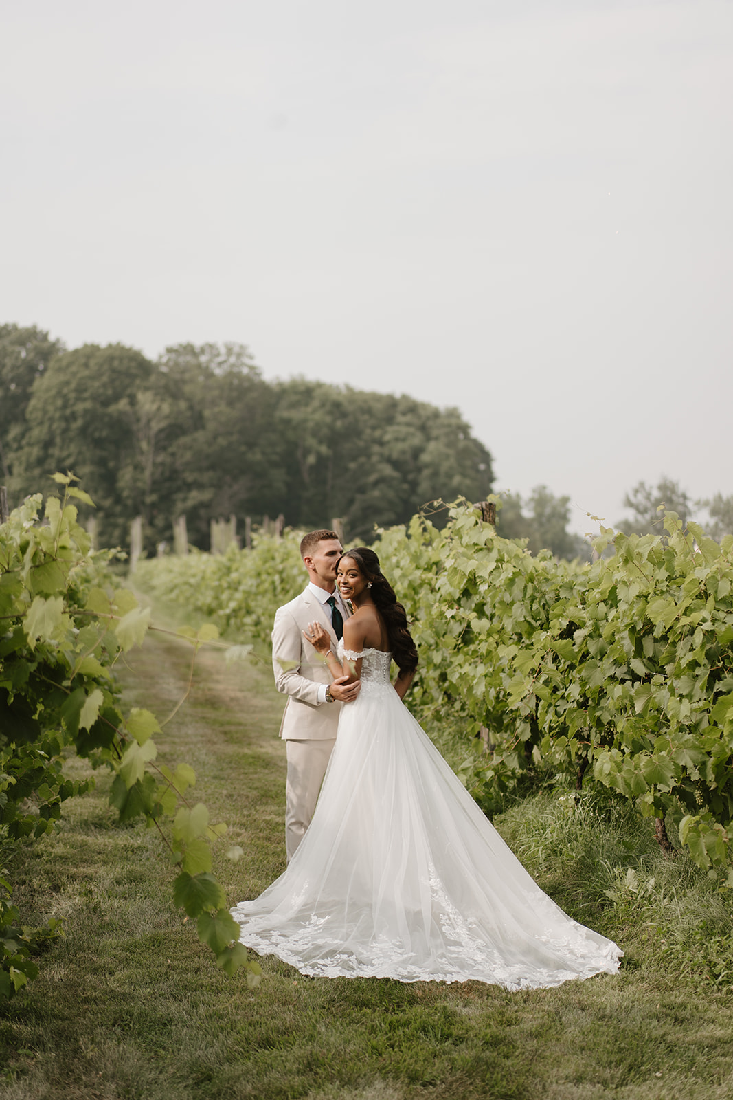 Bride and groom embrace between the vineyard rows on a soft summer day.