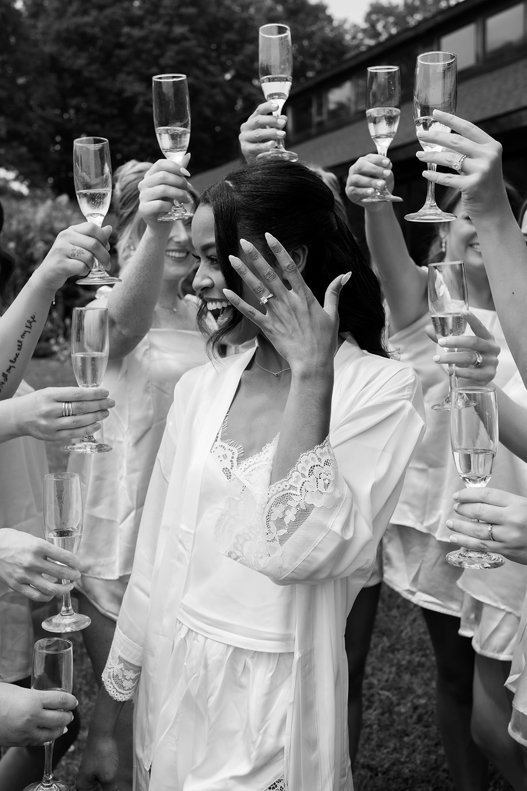 Black and white photo of the bride laughing as her bridesmaids raise champagne glasses around her.