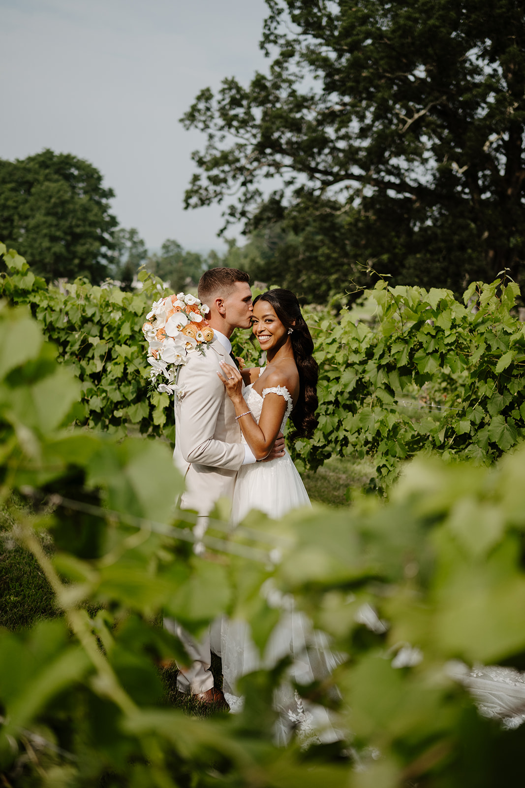 Bride and groom share a kiss in the vineyard, her bouquet lifted beside them.
