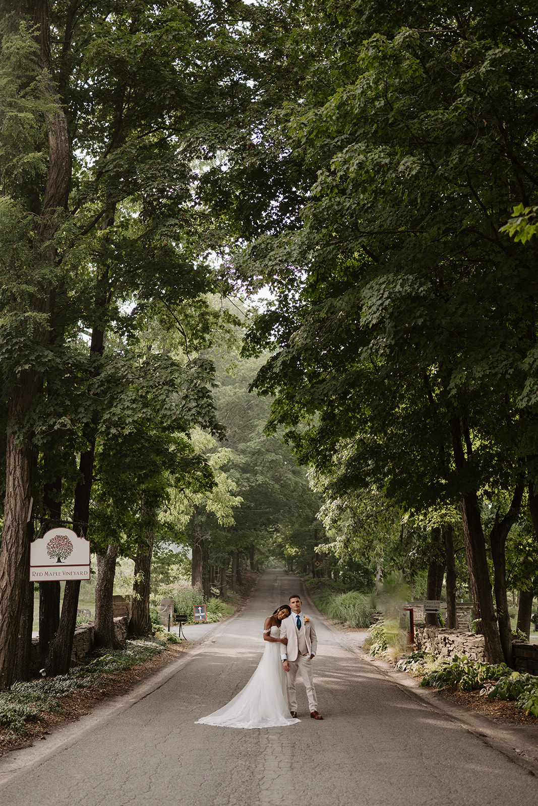Bride and groom standing on the tree-lined road beside the Red Maple Vineyard sign in West Park, NY