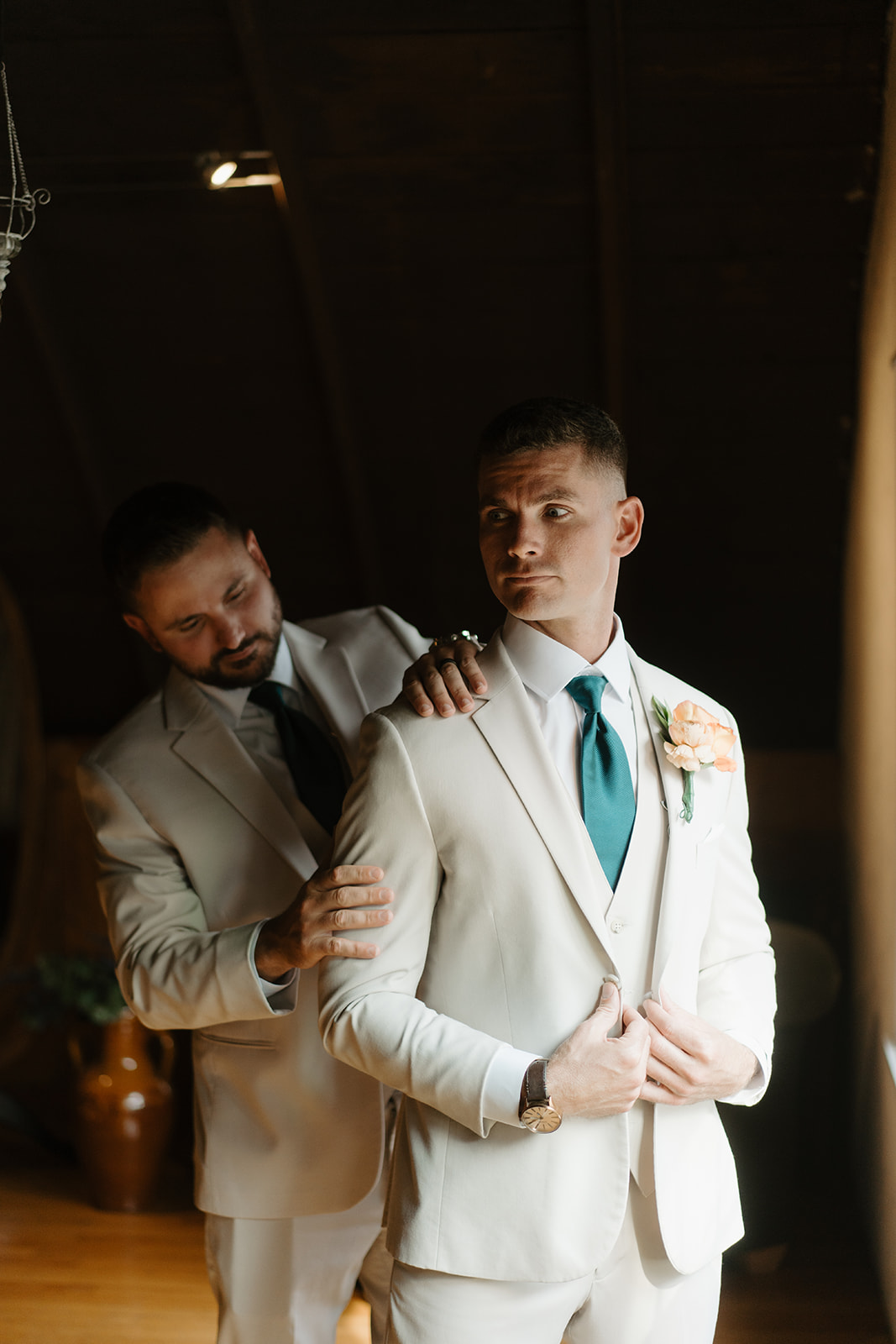 Groomsman rests a hand on the groom's shoulder as he buttons his jacket.