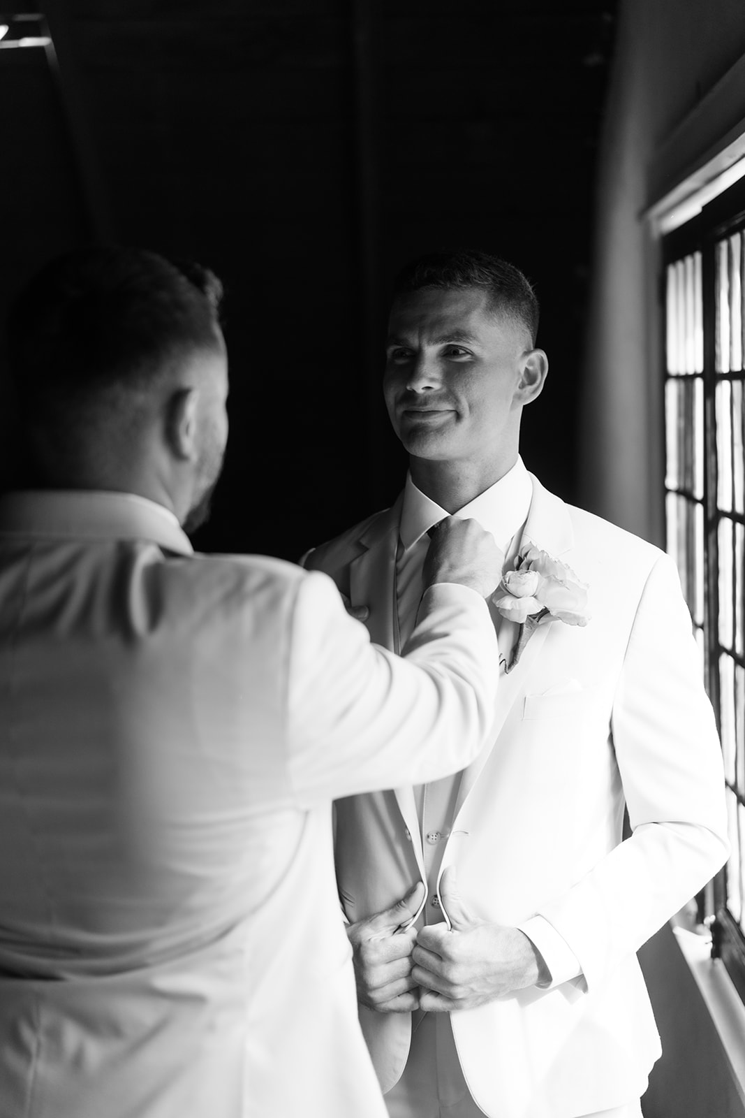 Black and white photo of a groomsman helping the groom with his jacket.