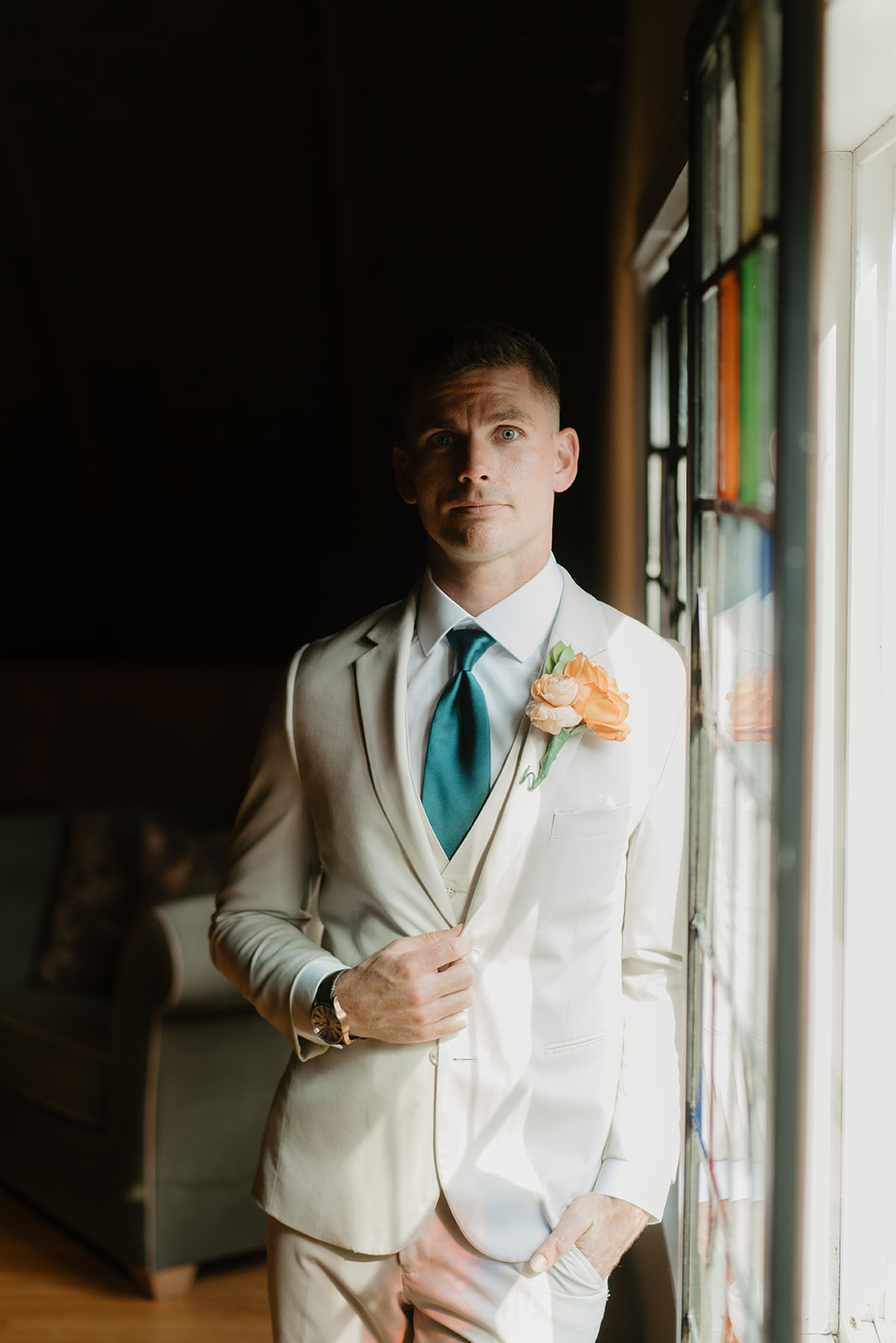 Groom in a cream suit and peach boutonniere stands beside the stained glass windows.