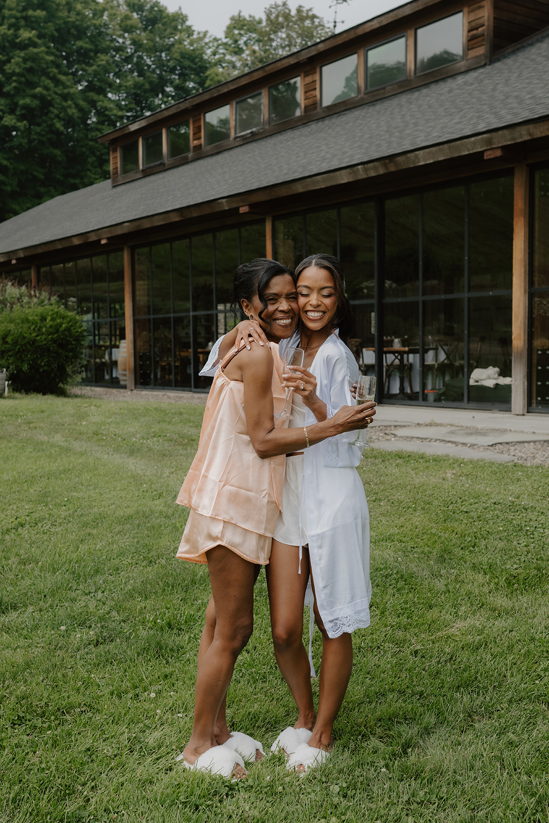 Bride and a bridesmaid in getting-ready pajamas toast champagne outside the glass-walled barn.