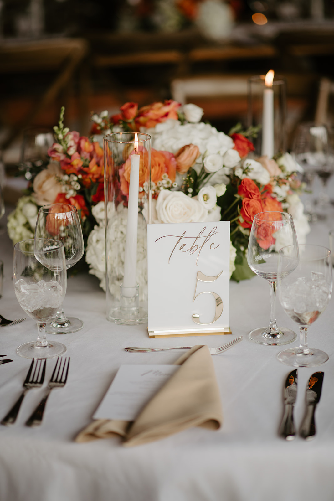 Close-up of a reception centrepiece with orange roses, candles, and a gold table number.