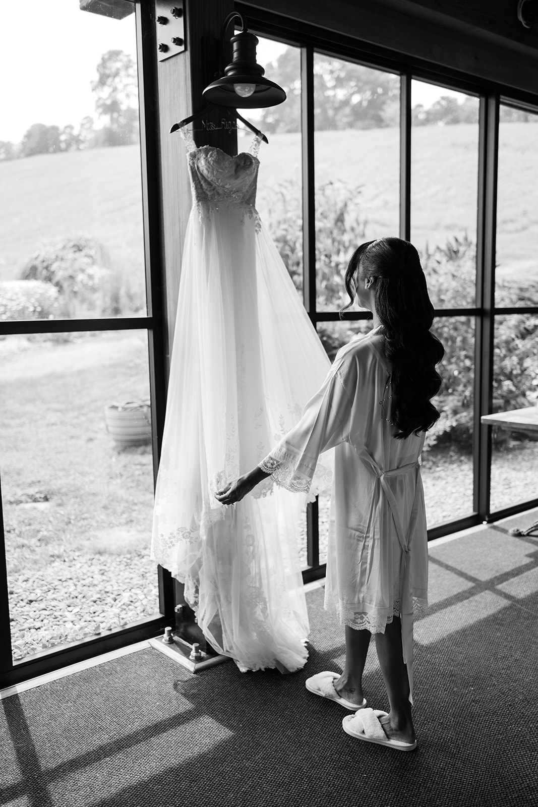 Black and white photo of the bride in her robe reaching for her gown hanging in a sunlit window.