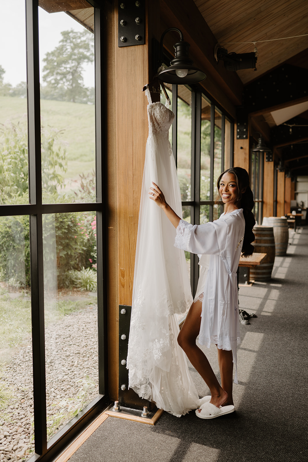 Bride in a white robe holds out her veil in a sunlit corridor of windows.